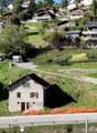 Aerial view of a hillside with several residential properties. Two-story houses with multiple windows. A grey detached house with balconies is under construction. Parking spaces are located near some of the houses. A pathway and a road with a guardrail ar