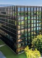 A modern, multi-story office building with a glass facade surrounded by lush greenery and trees. The building appears to be situated in a park-like setting with walking paths and landscaping visible in the foreground.