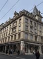 The image shows a large, ornate multi-story building with a prominent tower and decorative architectural features. The building has a Longchamp store on the ground floor and appears to be located on a busy city street with tram lines and pedestrians visib