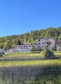A multi-story residential building with balconies, surrounded by trees and greenery in a scenic, hilly landscape.