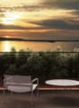 The image shows an outdoor patio or terrace overlooking a scenic lake or body of water. The patio has a round table and two chairs, providing a comfortable seating area to enjoy the beautiful sunset view over the water. The landscape is filled with lush g