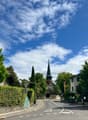 The image shows a picturesque street in a small town or village. The scene features a church steeple with a green roof, surrounded by lush green trees and hedges. The street is lined with buildings and has a parking area visible. The sky is blue with fluf