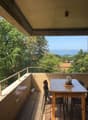 Covered balcony with wooden floor, glass railing, and a dining table with chairs overlooking a scenic view of trees and distant buildings