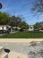 A large grassy area with trees and a paved walkway, surrounded by parked cars and buildings.