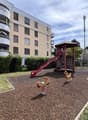 Playground with slide, climbing structure, and horsey ride toys, bordered by bushes, adjacent to a building with balconies.