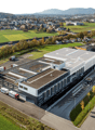 Large industrial building with multiple loading docks, surrounded by parking spaces and green fields. The building has a flat roof and is located in a suburban or rural area with mountains in the background.
