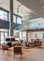 Large open-concept office space with concrete walls and ceiling, large windows, leather armchairs and sofas, and a spiral staircase leading to a mezzanine level.
