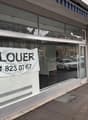 Shopfront with glass walls and doors, blue awning, white exterior, parked cars outside, signboard with a phone number and 'A LOUER' written in black