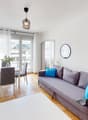 living room with dining area, gray couch, white table, chairs, white rug, white walls, wooden floor, white curtains, lamps, mirror, picture frame, television