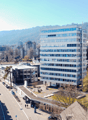 The image shows a cityscape with a mix of modern high-rise buildings and older, smaller structures. The city is surrounded by mountains and has a mix of residential and commercial areas. There are people walking on the streets and roads visible in the ima