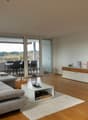 A modern and open living room with large windows and sliding glass doors leading to a balcony with an outdoor dining area and stunning views of the forest. The room features wooden floors, a comfortable grey sofa, a white coffee table, and decorative item