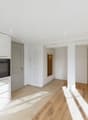 Open-concept living space with white walls, hardwood floors, built-in kitchen with oven and dishwasher, and a hallway leading to other rooms