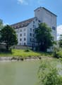 Large white building with multiple windows, situated next to a river, surrounded by trees and grass.