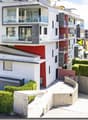 Modern apartment building with multiple floors, white and red facade, glass balconies, fenced entrance, hedges, and a small garden