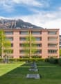 Modern apartment building with multiple floors, surrounded by greenery and a playground, with a view of the mountains