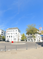 The image shows a street view of a city center. There are various buildings, including office buildings and shops, lining the street. The street has a roundabout in the center, and there are parked cars and pedestrians visible. The buildings have a mix of