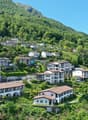 Hillside residential area with multi-story buildings, balconies, and parking spaces surrounded by lush green vegetation and mountains