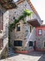 This image shows a traditional stone building with wooden shutters and balconies. The building has a rustic, Mediterranean-style architecture with a cobblestone pathway leading to the entrance. There are potted plants and a small bench visible, adding to