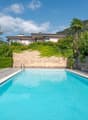 Two-story house with a swimming pool, surrounded by lush greenery and palm trees. The house has a balcony or terrace overlooking the pool area.