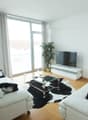 Spacious living room with large windows, white walls, hardwood floors, white leather sofas, glass coffee table, and a potted plant. The room has access to a balcony or terrace.