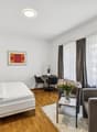Spacious bedroom with hardwood flooring, large windows with curtains, a double bed, a small round table with two chairs, and a vase of flowers on a side table.