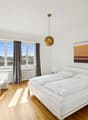 modern design bedroom with large windows, wooden floor, white walls, bed with pillows, white night stand lamps, white chair, wooden desk with picture frame