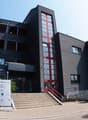 Three-story office building, black exterior, red accents, large windows, stairs leading to entrance, plants and rocks outside.