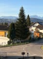 House with brown roof, yellow walls, balcony, solar panels, garage, driveway, trees, mountains in the background