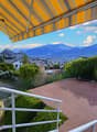 outdoor terrace with awning, railing, wooden floor, bushes, net, cityscape view, mountains in the distance
