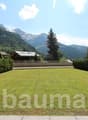 The image shows a scenic mountain landscape with a large grassy lawn in the foreground. There are several wooden chalets or cabins visible, with balconies or terraces. The mountains in the background are covered in lush green forests. The sky is blue with