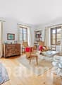 Living room with white walls, wooden floor, beige rug, large window, wooden cabinet, couch, books on shelves, coffee table, lamp