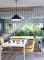 Open-concept living and dining area with large windows overlooking a garden, hardwood floors, black metal and wood staircase, white dining table with yellow chairs, black pendant light fixture