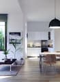 Spacious open-plan living area with large windows, modern gray sofa, wooden dining table and chairs, kitchen with white cabinets, and a black pendant light fixture.