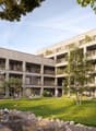 Multi-story apartment building with balconies, surrounded by trees and greenery, with a landscaped courtyard and children playing in the foreground.