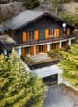 2-story wooden chalet-style house with a slanted dark roof, large windows, and a balcony/terrace. The house is surrounded by lush green pine trees and is situated in a forested, mountainous area.