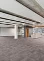 Large open office space with exposed concrete ceiling and beams, gray carpeted flooring, and several windows along the walls. The space appears to be a commercial or office environment, with a clean and modern design.