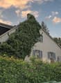 Two-story house with a gabled roof, surrounded by lush greenery and vegetation. The house has shuttered windows and a balcony or terrace visible on the upper level. The exterior walls are painted a light color, and the roof appears to be made of tiles or