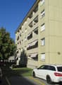A multi-story building with beige walls and brown trims. It has awnings on the balconies. There is a tree and a play area nearby. There are 3 cars parked in front of the building.