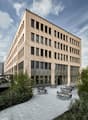 Multi-story office building with a modern, rectangular design featuring large windows and a paved outdoor seating area with tables and chairs.