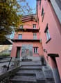 A two-story pink building with balconies, surrounded by trees. The building has a mural painting on the front and a set of stairs leading to the entrance.