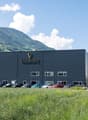 Large black industrial-style building with the Valmont logo, surrounded by a grassy area and parked cars in the foreground. The building appears to be a commercial or industrial facility located in a mountainous, forested area.