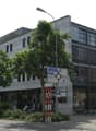 Multi-story office building, white exterior, glass windows, ground-level storefronts, trees and bushes in front, cars parked on street, street light in front.