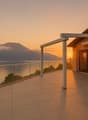 Two-story house with a balcony/terrace overlooking a scenic lake and mountains at sunset. The house has a tiled roof and large windows, with a glass railing on the balcony providing an unobstructed view of the beautiful natural landscape.