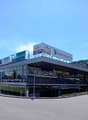 Commercial building with glass facade, multiple signs, parking lot, entrance with stairs, street lights, greenery