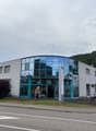 A modern, multi-story commercial building with a glass facade and a parking area in front of it. The building appears to be located near a road or highway.