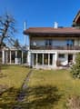 2-story house, brown roof, large glass windows, balcony, garden, potted plants, pathway, grassy lawn, trees, nearby houses