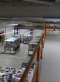 The image shows a large industrial facility or warehouse. The space is filled with various equipment, machinery, and storage racks. There are several workstations and production lines visible. The ceiling is high and the lighting is industrial. The overal