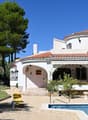 Two-story Mediterranean-style villa with a red tile roof, white exterior walls, and a tower feature. The house is surrounded by lush greenery, including palm trees and other vegetation. There is a swimming pool in the foreground with a patio and outdoor s