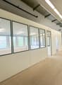The image shows a large open office space with white walls, windows, and a concrete floor. The space appears to be unfinished, with exposed ceiling beams and lighting fixtures. The room has multiple glass-paneled walls, creating separate sections or offic