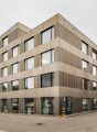 A multi-story office building with a modern, angular design. The exterior is made of beige and brown panels, with large windows throughout. There is a parking area visible in the foreground of the image.